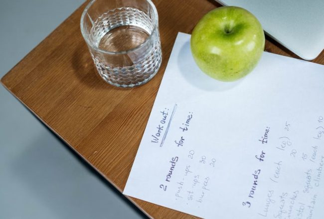 A green apple, glass of water, and workout plan on a wooden desk symbolize healthy living.