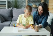 Small boy is drawing with markers together with caring mother in cozy room at home enjoying activity making picture. Creative kids and loving parents concept.