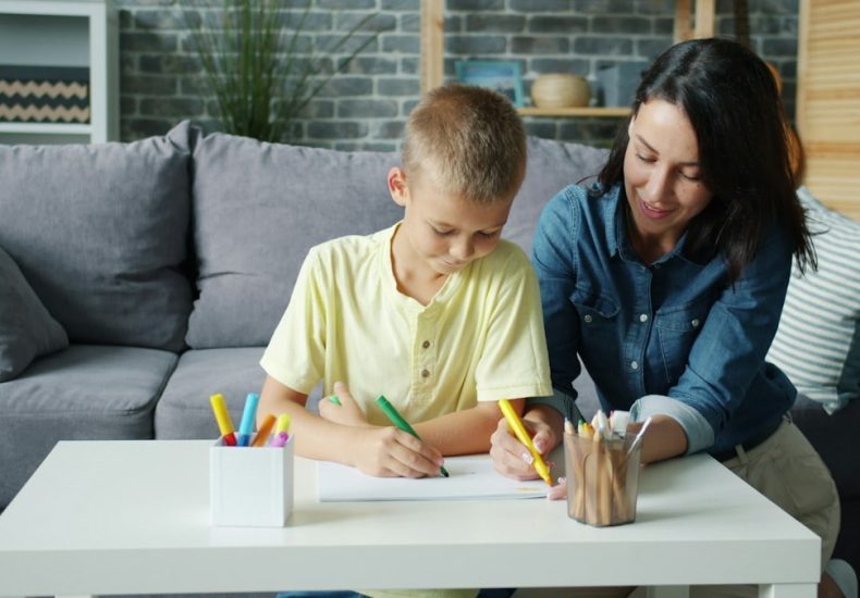 Small boy is drawing with markers together with caring mother in cozy room at home enjoying activity making picture. Creative kids and loving parents concept.